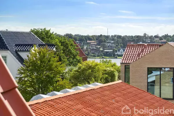 Røde tegltage og grønne træer ses tydeligt, med en udsigt over en bydel og en sø i baggrunden under en klar blå himmel.