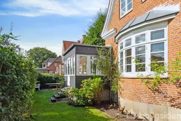 Murstenshus med hvide vinduer har en tilbygning i glas. Det står i en velplejet have med græsplæne og blomsterbede. Andre huse ses i baggrunden under en blå himmel.