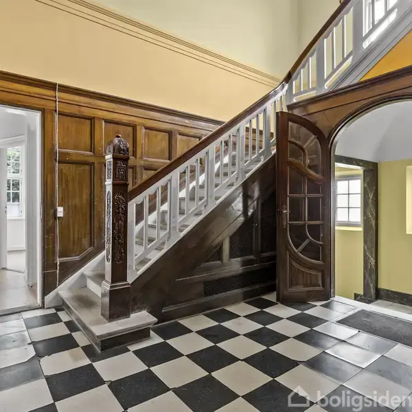 Trappeopgang med mørkt trægelænder, fører op i en lysere etage. Omkring er der hvide døre og brune vægge. Gulvet er sort-hvid ternet, og lys strømmer ind gennem vinduerne.