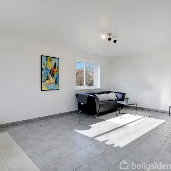 En stue med en sort sofa står mod en hvid væg. Rummet har et farverigt maleri og et vindue, der lader sollys komme ind. Fliser dækker gulvet.