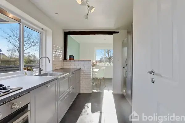 Lyst køkken med stålskabe og vask, åbner mod en spisestue med spisebord og stole, store vinduer giver naturligt lys, udsigt til udendørsareal med træer.