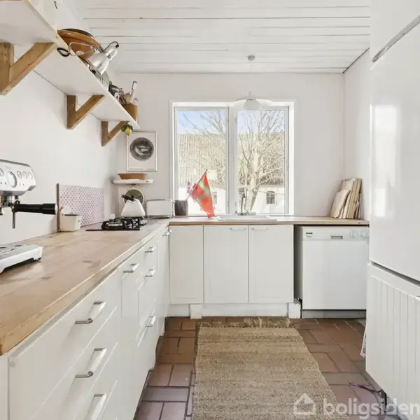 Køkken med hvidmalede skabe og træbordplader rummer en espressomaskine til venstre. Gennem vinduet ses et træ og et dansk flag. Klinkegulv og hylder med køkkengrej skaber hjemlig atmosfære.