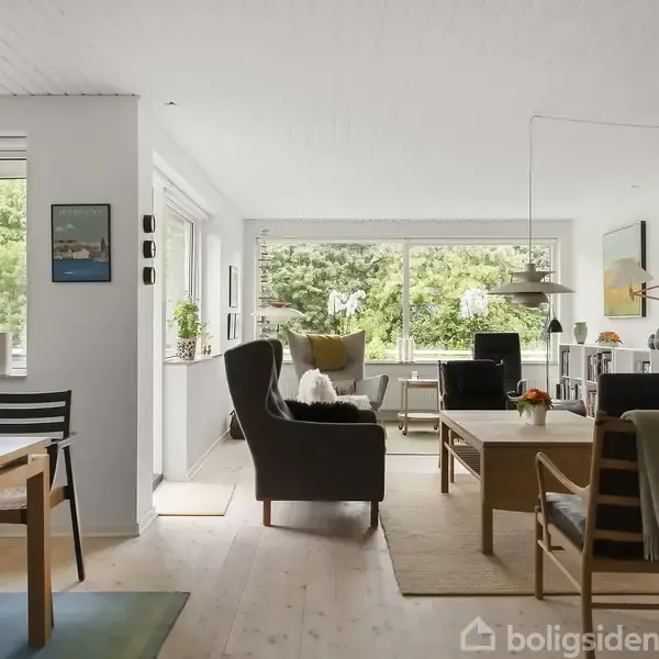 Stue med moderne møbler står i et lyst rum. Store vinduer viser udsigt til en grøn have. Et spisebord med stole er placeret til venstre.