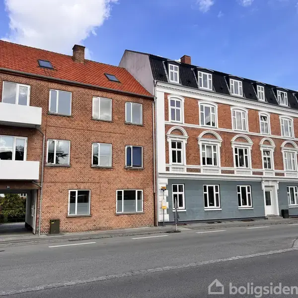 En række af to bygninger med røde mursten. De står stille langs en gade med gråt fortov under en blå himmel og nogle skyer.