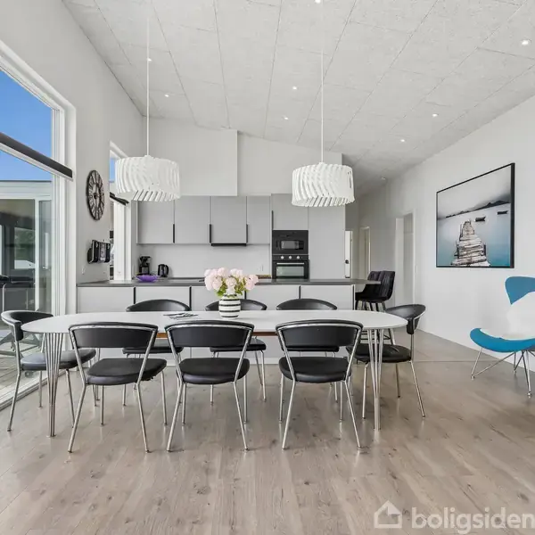 Spisebord med otte stole står i en lys stue med trælaminatgulv. Store vinduer giver udsigt til terrasse. Køkkenet har moderne apparater, og to pendellamper hænger over bordet.