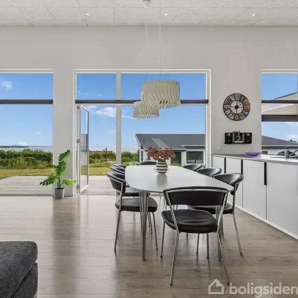 Moderne spisestue med sort-hvidt farveskema, hvor et ovalt bord med stole står centralt. En glasdør åbner ud mod en terrasse og have med blåt himmeludsigt.