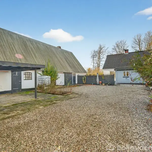 En grusbelagt gårdsplads ligger mellem to hvidmalede bygninger med skrå tage. Til venstre ses en bueformet dør og små vinduer, omgivet af træer og buske under en blå himmel.