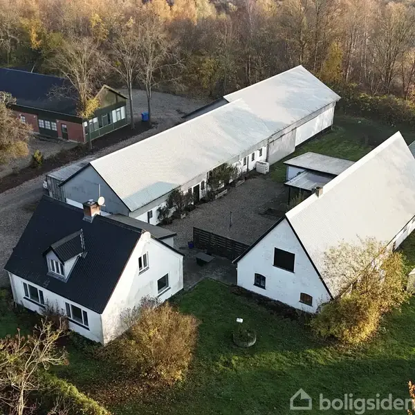 Et hvidt hus med sort tag ligger i et landligt område omgivet af træer. Bagved er der flere aflange bygninger med lyse tage og en gårdsplads.