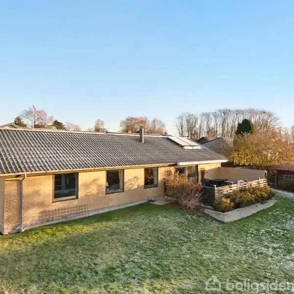 Et enfamiliehus af mursten står stille i en velholdt have med frost på græsset i et villakvarter med nøgne træer i baggrunden.