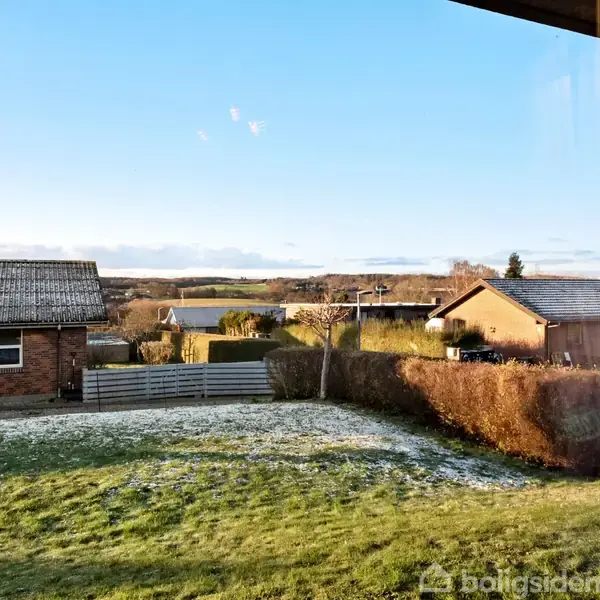 Udsigt gennem et vindue viser en græsplæne med frost i en baghave. Et hegn og to huse omkranser området. Himlen er klar og blå.