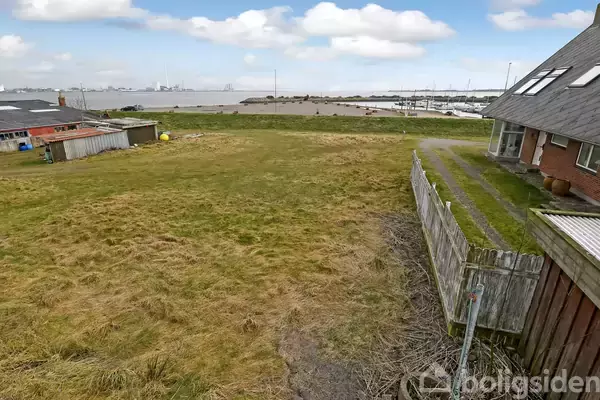 Græsplæne strækker sig ved siden af et hus med tagrytter, omgivet af hegn. I baggrunden ses stranden og havet, med en klar, blå himmel og spredte, hvide skyer.