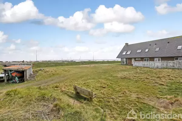 Hus med skråt tag står stille i en åben mark, omgivet af grønne græsplæner og med en trækonstruktion tæt på. Baggrunden viser en klar himmel med spredte skyer.