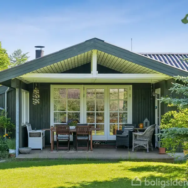 Et træhus har en veranda med hvide møbler og spisestole. Det ligger i en frodig have, omkranset af grønne buske og træer under en klar blå himmel.
