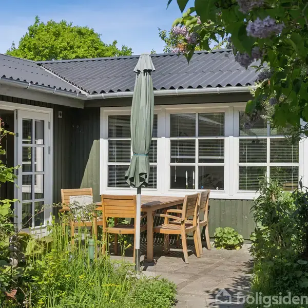 Terrassebord med træstole står under en parasol i en grøn have. Et hus med grønne vægge og vinduer omgiver terrassen, omgivet af frodig vegetation og klar blå himmel.