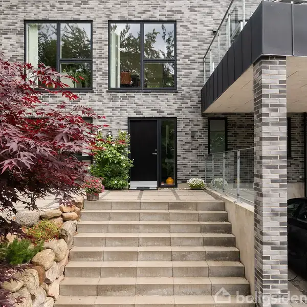 Husindgang i moderne stil med muret facade i grå mursten. Sorte døre og vinduer. Trappe fører op til indgang, omkranset af planter i små bede og en åben carport på højre side.
