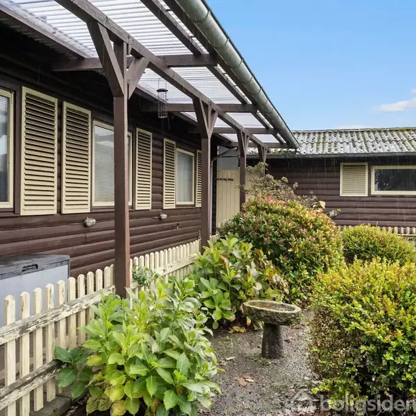 Brun træbygning med lukkede skodder står stille i en have med buske og blomster, omkranset af et lavt, hvidt stakit. Et transparent tag beskytter en del af facaden.