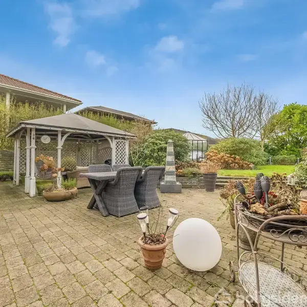 En overdækket pavillon med et spisebord og stole står på en flisebelagt terrasse omgivet af planter og potter. I baggrunden ses græs, træer og nabohuse under en blå himmel.