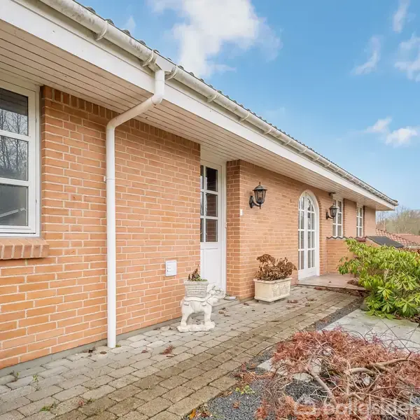 Rødstenshus med hvidt tagrender, en sti af fliser langs facaden, krukker med planter foran døren. Omgivet af blå himmel og buske, præget af en afslappet atmosfære.