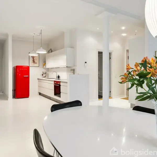 Rundt spisebord med en vase af orange blomster placeret i et lyst, moderne køkken. Et iøjnefaldende rødt køleskab står i baggrunden, omgivet af hvide vægge og lamper.