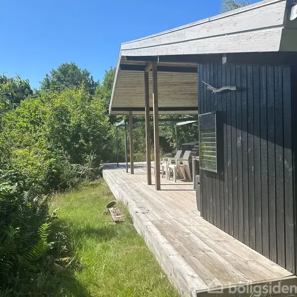 En sort træbygning med terrasse skyggefulgt af et halvtag. Flere hvide stole står under taget. Omgivet af frodig grøn vegetation og græsplæne under en klar blå himmel.