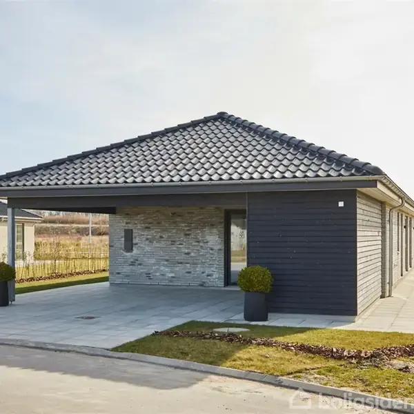 En moderne bolig med gråt tegltag står ved en flisebelagt indkørsel. To runde buske er placeret i krukker ved indgangen. Den omgivende natur består af åbne marker.