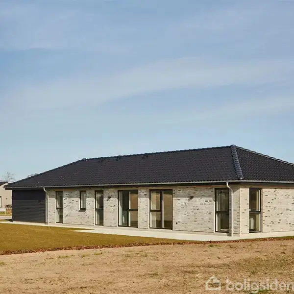 En moderne, énetages bungalow med mørkt tag står på en græsplæne. En flagstang med "MILTON" svajer i vinden. Rækkehuse kan ses i baggrunden under en blå himmel.