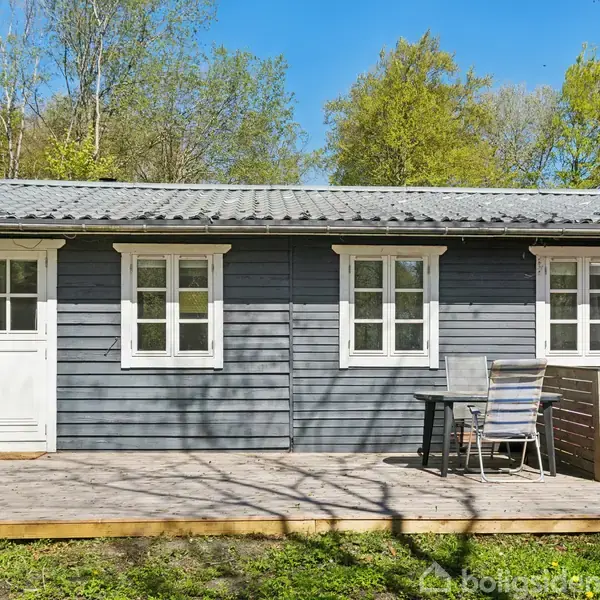 Et gråt træhus står stille med et lyst tag i en solrig have. Foran huset er en terrasse med bord og stole. Træer omkranser området.