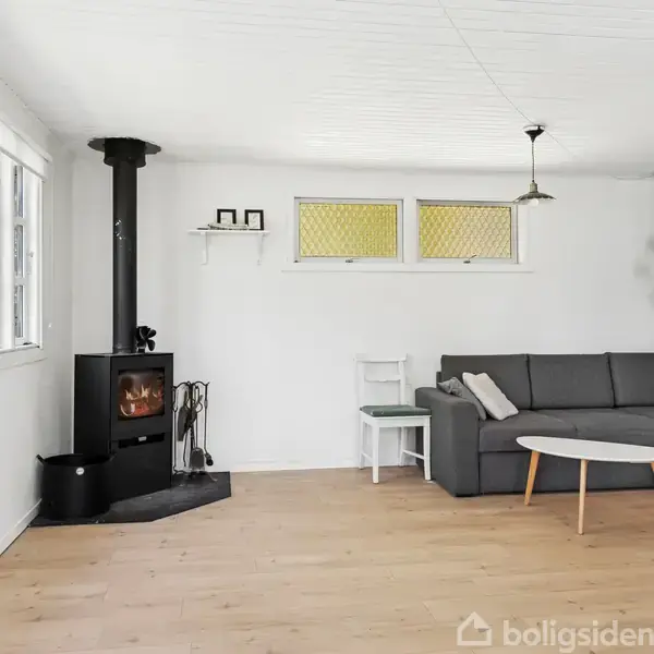 Brændeovn opvarmer hjørne af stue med hvide vægge og parketgulv. L-formet sofa og rundt sofabord står ved grøn væg. Vinduer med gult glas slipper lys ind.