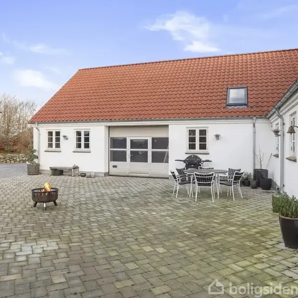 Et hvidt hus med rødt tag står på en stor, velplejet græsplæne. I baggrunden ses nabohuse og træer under en klar, blå himmel.