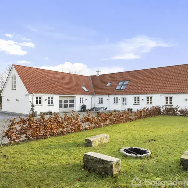 Hvidt hus med rødt tegltag; udendørs spisebord med stole står på en flisebelagt gårdsplads; bålplads til venstre; omgivet af træer og blå himmel.