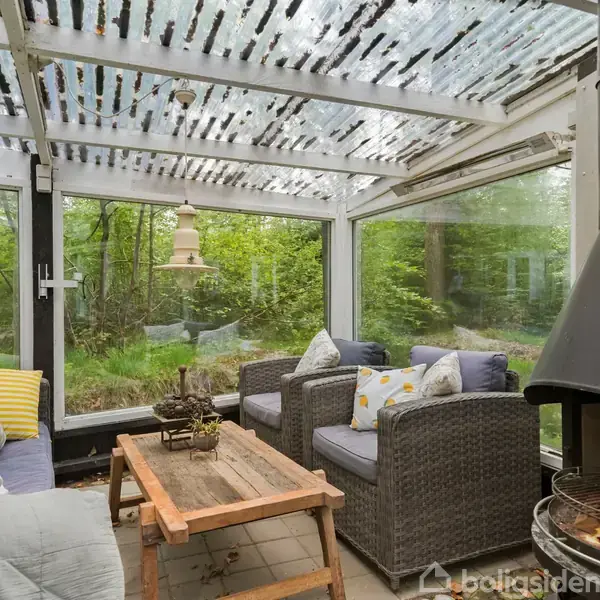 Et overdækket terrasseområde med hvide rammer, hvor rattanmøbler er placeret omkring et træbord. En åben pejs står til højre, omgivet af grøn natur udenfor glasset.
