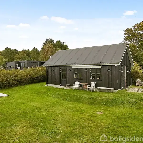 Et sort træhus står stille på en grøn græsplæne omgivet af buske og træer. To havestole og et bord står foran huset. Skyer ses på en blå himmel.