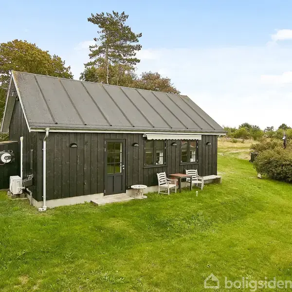 En sort træbygning med skråt tag står i en grøn have. To stole og et bord foran huset. Omgivelserne inkluderer buske og træer under en blå himmel.
