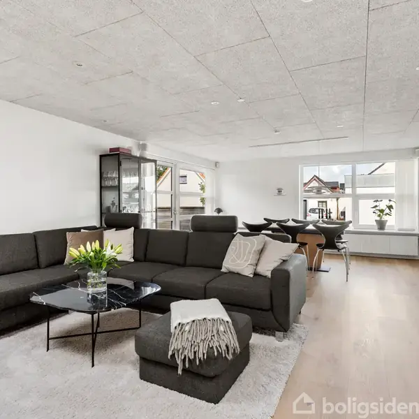 En mørkegrå hjørnesofa står i centrum af en stue på et lyst trægulv. Et sofabord med blomster, tæppe og puder skaber en indbydende atmosfære, mens store vinduer slipper lys ind.