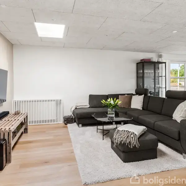 En stue med en stor mørkegrå hjørnesofa fyldt med puder, placeret på et lyst tæppe. Et sofabord med en blomsterbuket foran sofaen. Et fladskærms-TV hænger på væggen. Bagved ses en skænk med glaslåger. Gulv i lyst træ, store vinduer med udsigt.