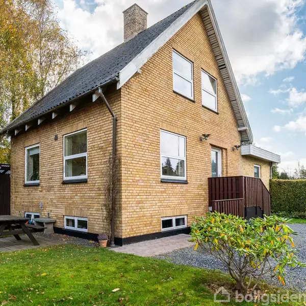 Et to-etagers gule murstenshus står i en grøn have med en grussti. Borde-bænkesæt er placeret til venstre, og træer er synlige i baggrunden under en delvist skyet himmel.