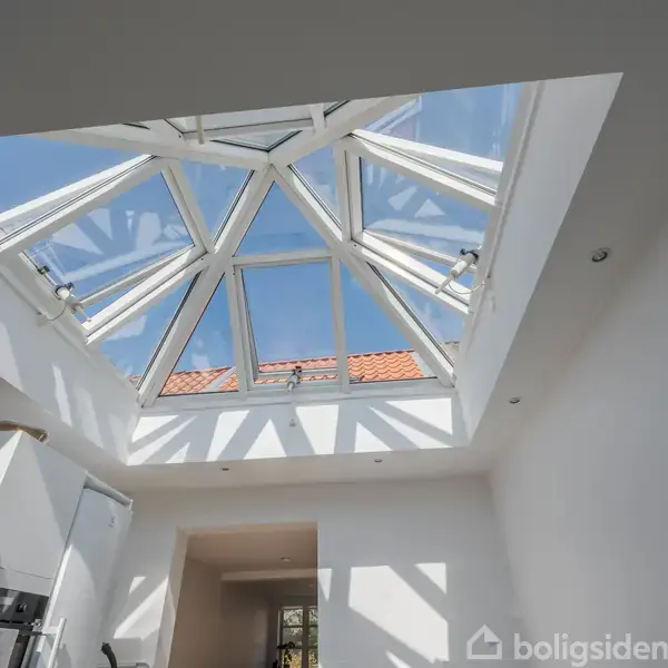 Glasloft dominerer rummet, slipper sollys ind gennem trekanter. Det hvide interiør inkluderer hvide vægge og køkkenskabe. Rummet er lyst med skarpe skygger og en klar blå himmel udenfor.