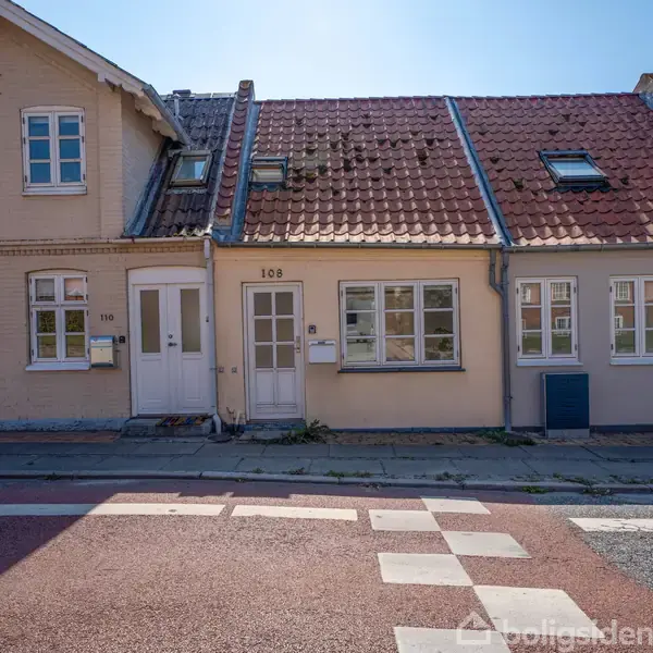 En rækkehusbygning med rødt tegltag og lyse vægge står langs en stille gade. Husnumrene 108 og 106 er synlige ved indgangsdørene. Foran er en fortov og vej.