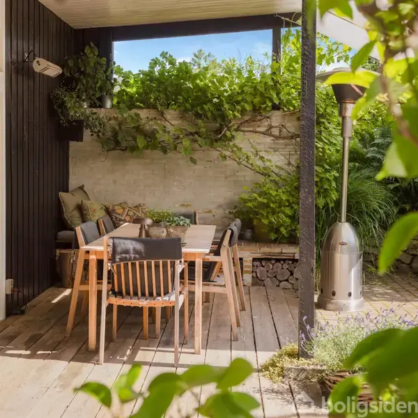 Spisebord med fire stole står på en træterrasse. Bredde vinduer og grønne planter skaber en hyggelig atmosfære. En terrassevarmer står ved siden af bordet, omgivet af frodig vegetation.