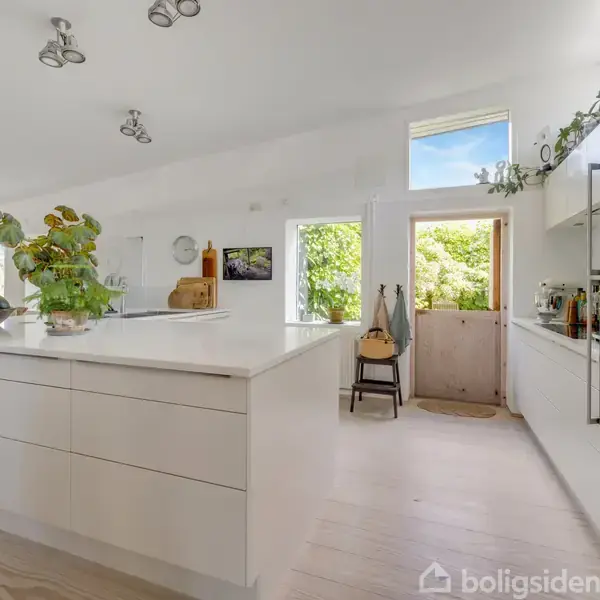 Et lyst og moderne køkken har en stor hvid køkkenø med planter. Rummet er åbent med trægulv, store vinduer og en dør, der fører ud til en grøn have.