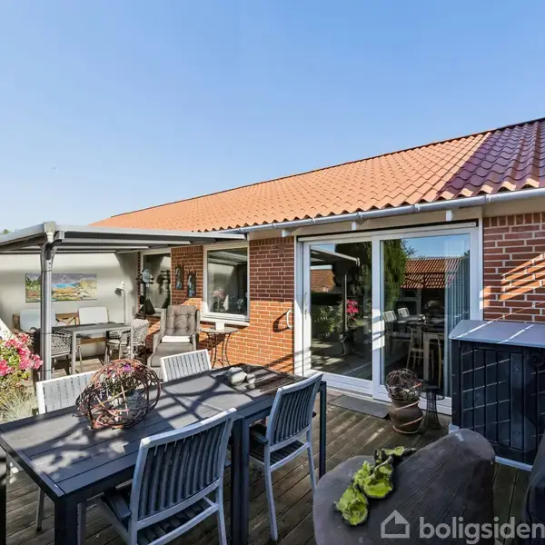 Terrasse med spisebord og stole, omgivet af træfliser, blomsterkrukker og buske. Til venstre ses en åben dør til huset, som er bygget i røde mursten med rødt tag.