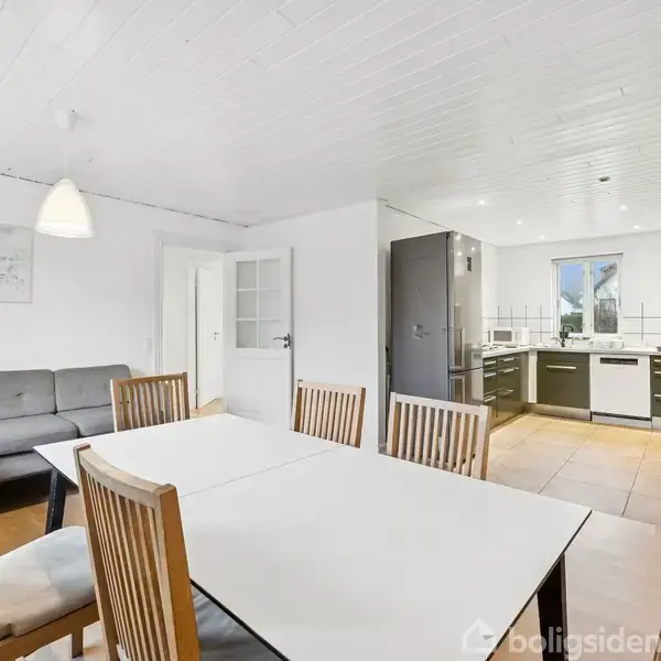 Et spisebord med træstole står midt i et lyst rum. Til venstre er en grå sofa, og bagved ses et moderne køkken med hvide og grønne elementer.