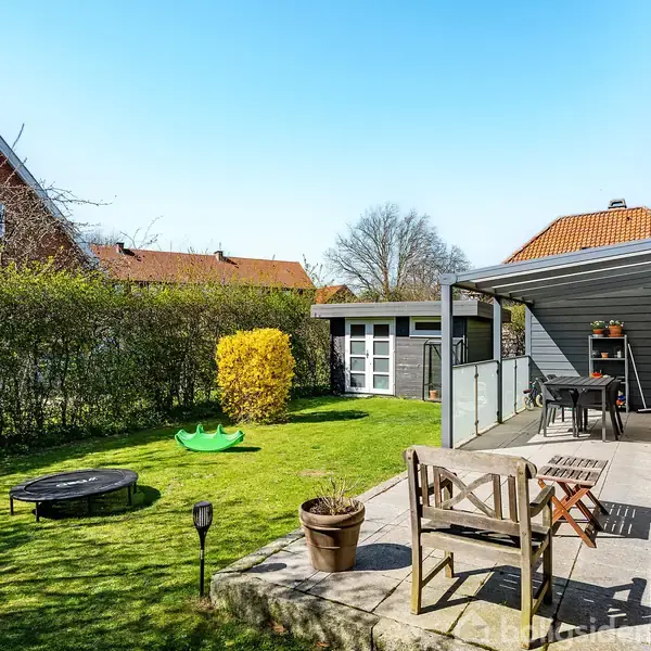 Terrasse med træmøbler står under en halvt overdækket veranda. En grøn have med en trampolin og legetøj omgiver området, mens nabohuse ses i baggrunden.