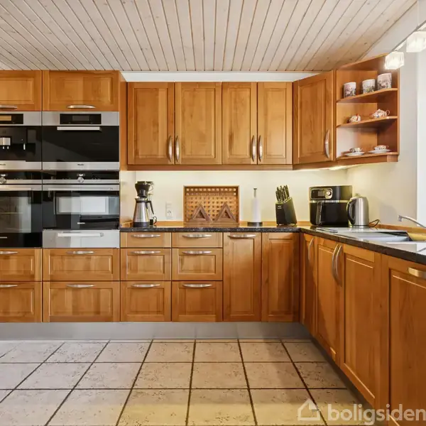 Køkken med træskabe og moderne apparater, såsom ovne og en kaffemaskine, omrammet af lyse fliser og et stort vindue til højre, som lader lys strømme ind.