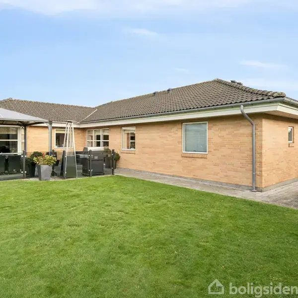 Huset er en enkelt-etagers bygning med rød mursten og mørkt tag. Det står fredeligt i en velholdt have med grøn græsplæne og terrasseområde.