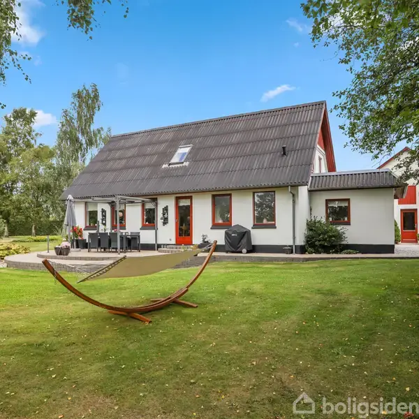 Et hus med skråtag står stille i en rummelig have med grønne træer og en hængekøje. Haven er pænt anlagt med en terrasse og en græsplæne.