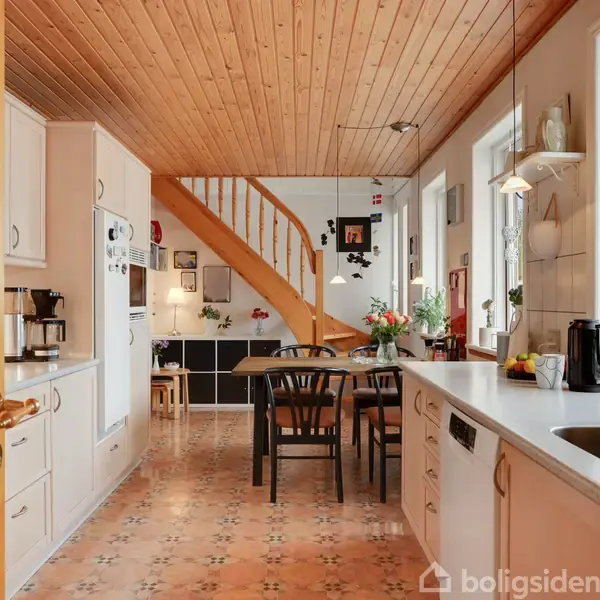 Et køkken med et langt hvidt bord og vask, sort spisebord med stole. Trappen fører til en anden etage. Rummet er lyst med træloft og vinduer.