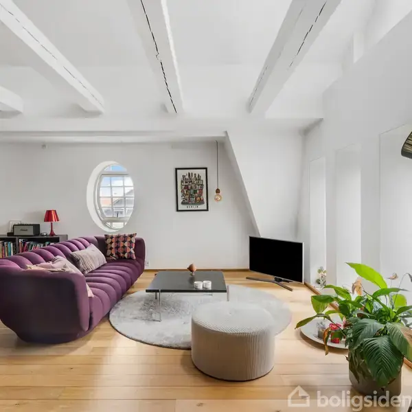 En lilla sofa står placeret i en lys stue med trægulve. Ovale vinduer og en gulvlampe skaber en hyggelig atmosfære. En plante og TV er placeret på hver sin side.