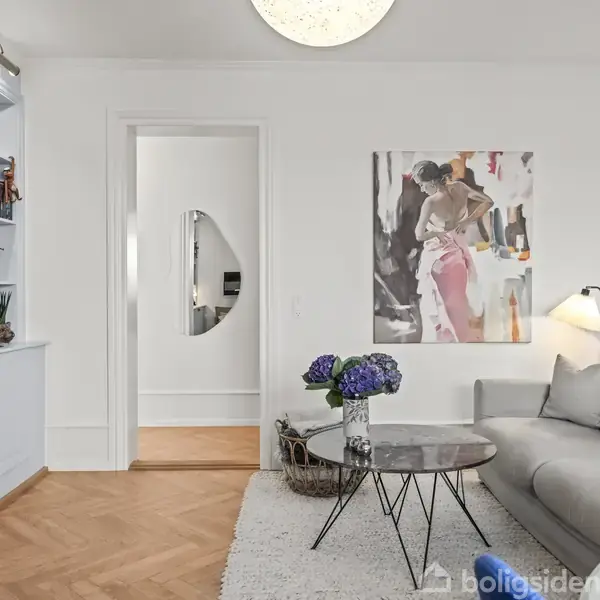 Stue med grå sofa og runde borde, dekoreret med lilla blomster. En farverig kunstplakat hænger på væggen. Til venstre står en hvid bogreol med bøger og dekorationer.