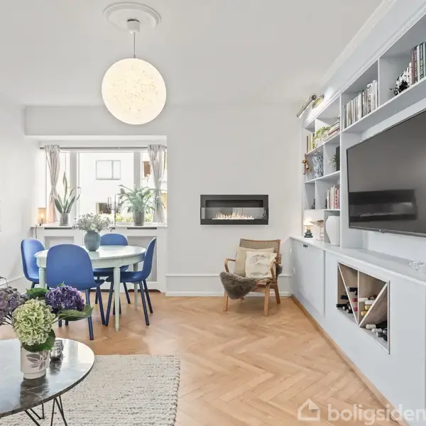 Stue med sofa og sofabord, udsmykket med blomster. Rundt spisebord med blå stole ved vinduet. Indbygget reol med TV og bøger på hvid væg. Pejs ved siden af stol.